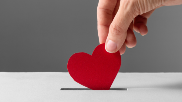 A hand delivers a bright red paper heart into a donation box.