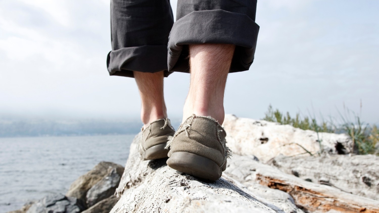 A person wearing casual shoes balances on a rock near a lake.