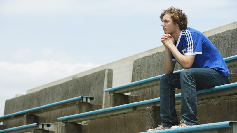 A young man sits on a bench under a clrear sky and looks out.