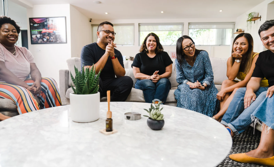 Six adults in colorful clothing laugh together on a soft gray sofa around a white marble table with textured green plants.
